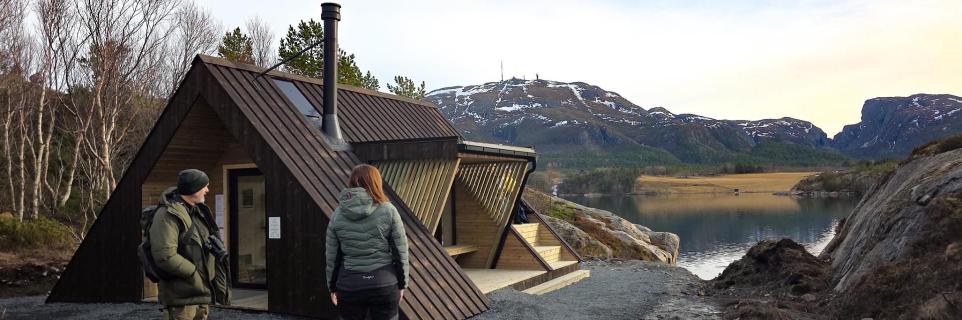 To personer iført turklær står ved en trekantformet hytte, og ser ut mot havet. 