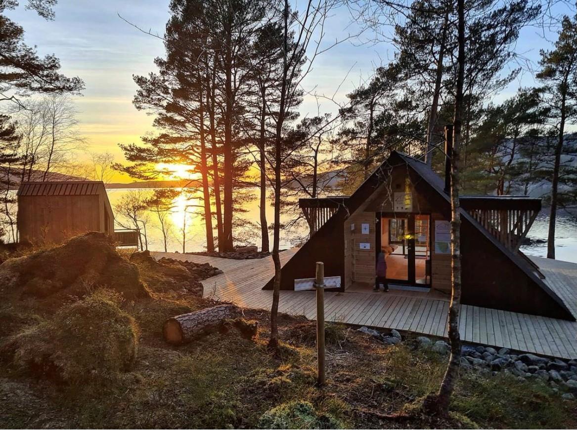 En hytte formet som en trekant står i en skog meg utsikt til et vann/fjord.