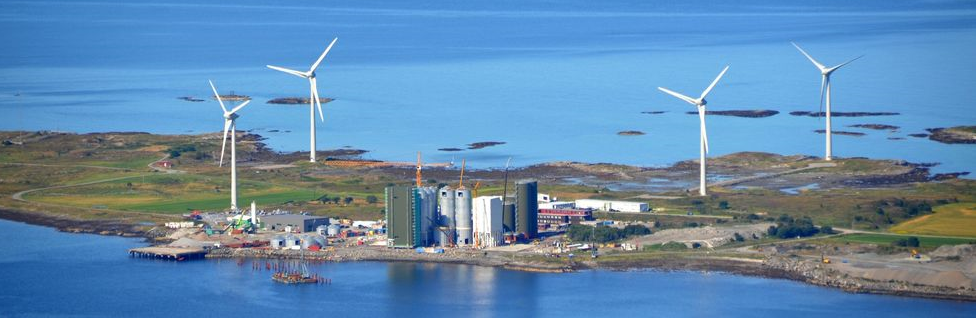 Overblikksbilde av av et industriområde med vindmøller og ulike bygg. Plassert rett ved havet. - Klikk for stort bilde