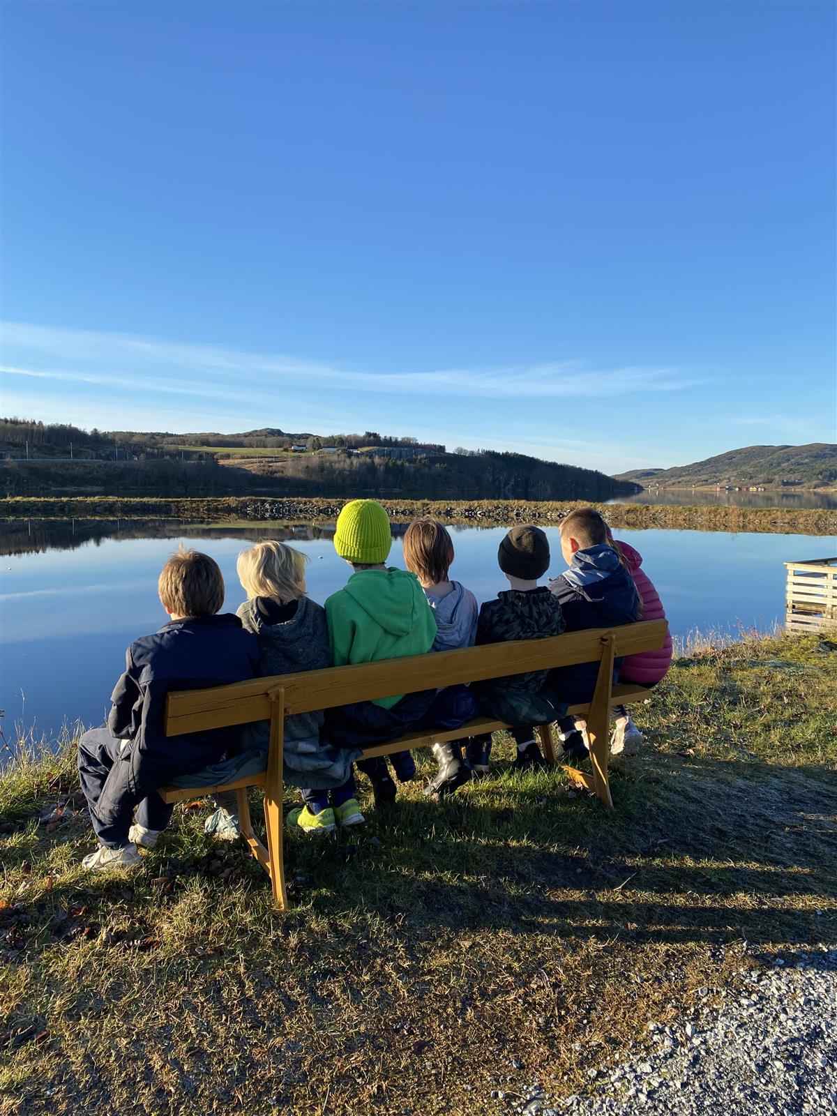 Viser 6 personer som sitter på en benk ved et vatn, skog og blå himmel i bakgrunn - Klikk for stort bilde