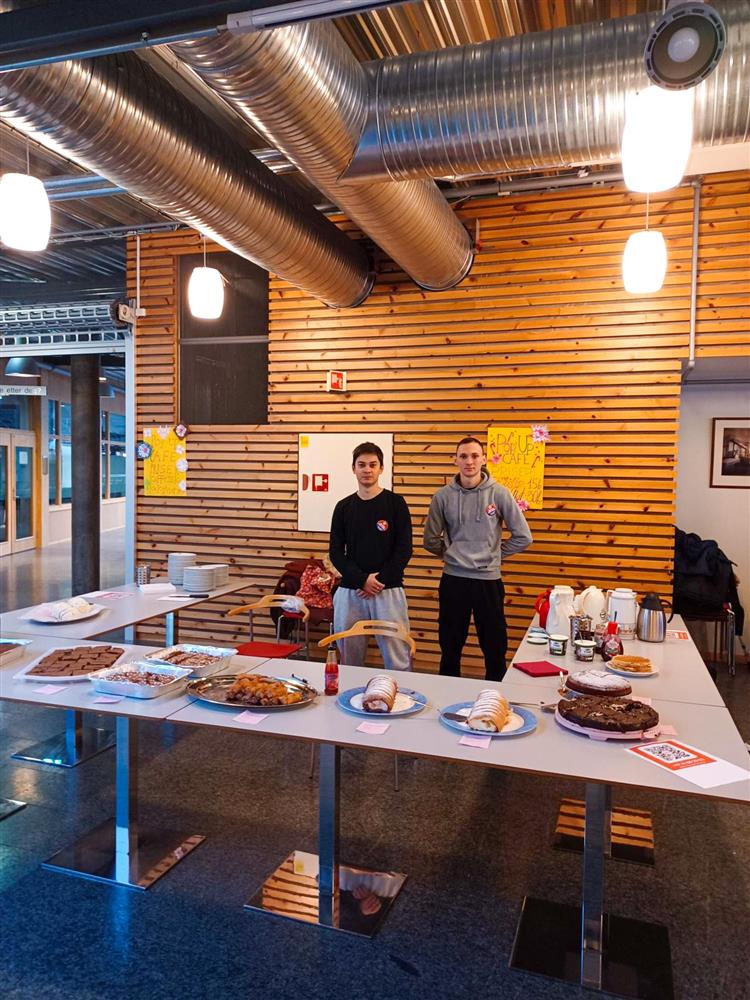 To unge menn som står bak et hesteskoformet bord med kaker og mat på.  - Klikk for stort bilde