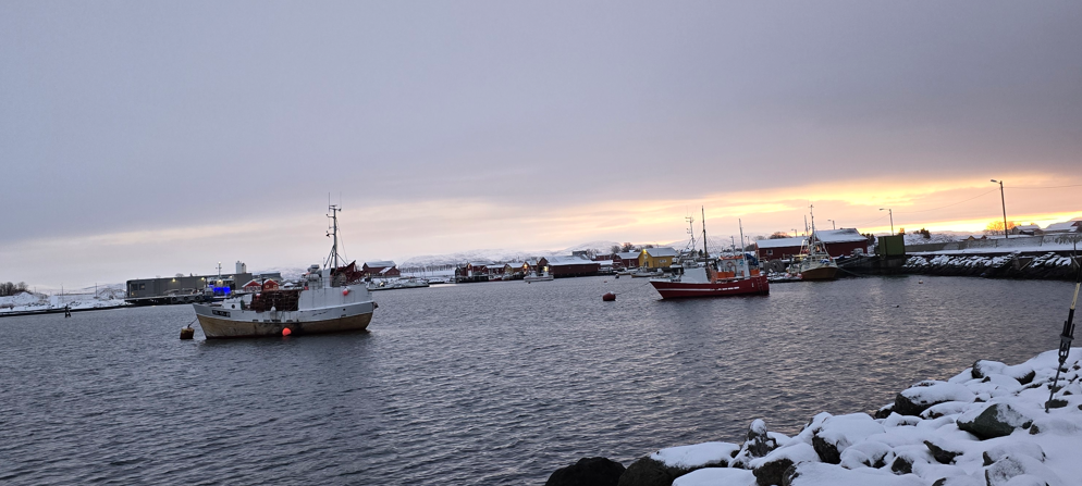 Bilde av båter ved et Skipsleie med solnedgang i bakgrunnen. Himmelen er overskyet og et tynt lag med snø dekker steinene ved havet. - Klikk for stort bilde
