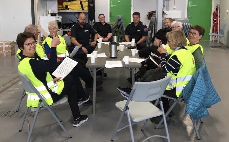 Sanitetsdamer med gul vest i samtale med folk fra brannvesenet over en kopp kaffe..  