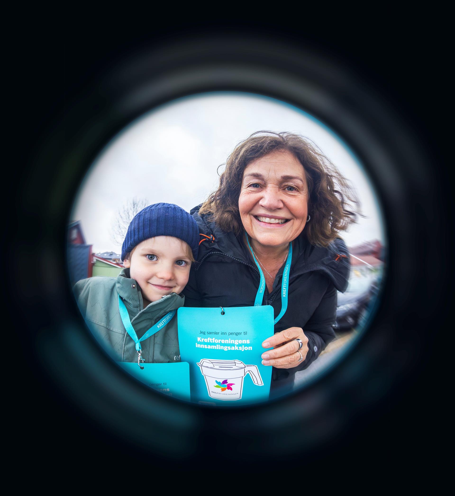 En voksen og et barn smiler mot kamera og holder opp lapper som viser at de er fra Kreftforeningens innsamlingsaksjon. Lappene er festet på et turkis bånd som henger rundt halsene deres. Bildet er tatt gjennom et dørkikkhull, som gir en mørk, rund ramme rundt motivet. 