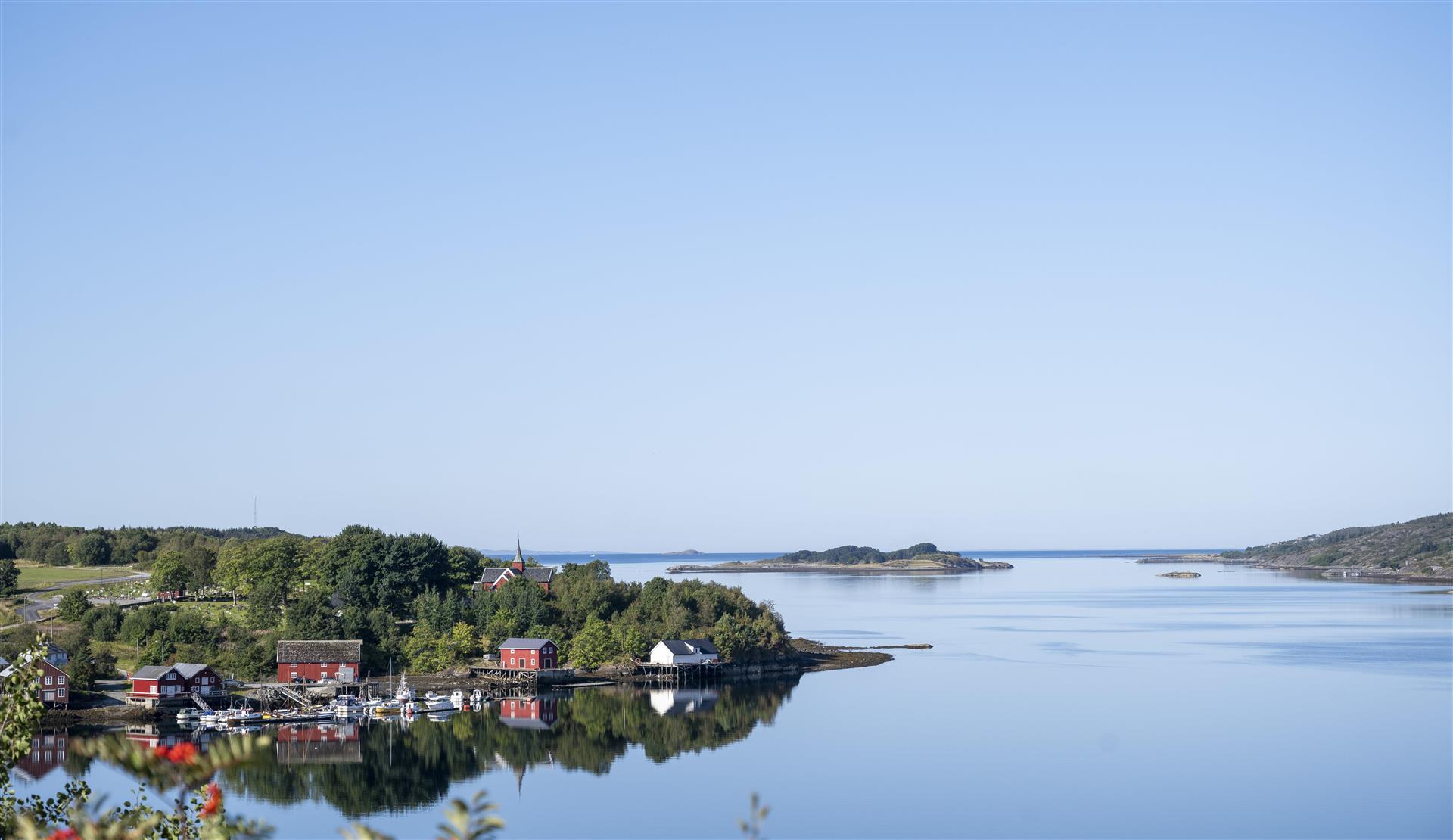 Utsikt ut fra kysten. Landskapsbilde med hav og himmel. Til venstre ser man ei kai med båter og rødebygg langs kystlinjen. Rød kirke i bakgrunnen. 