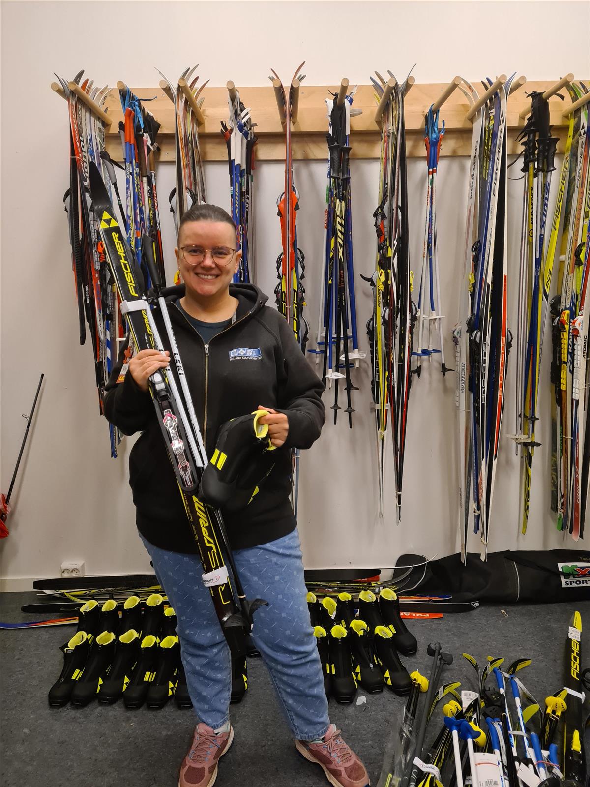 Bildet viser en dame med ski, skisko og staver med masse skiutstyr i bakgrunnen - Klikk for stort bilde